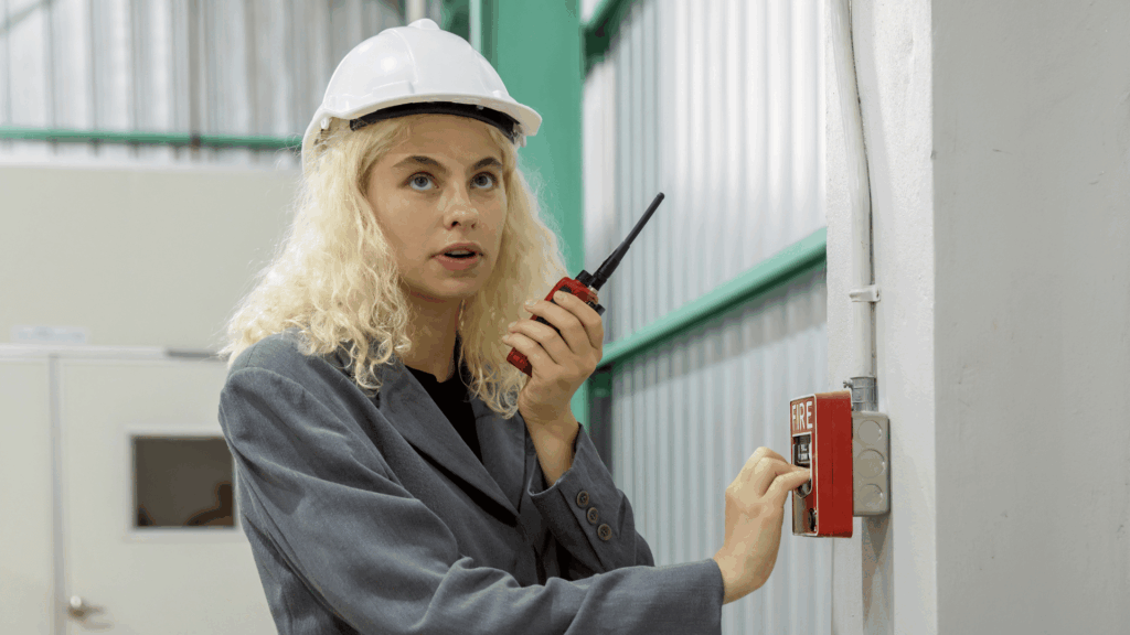 Femme dans un entrepôt, muni d'un casque sur la tête, et d'un talkie-walkie dans la main gauche, qui déclenche l'alerte incendie de la main droite